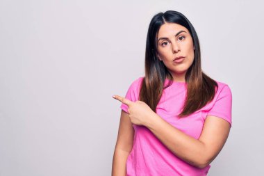 Genç, güzel, esmer, sıradan pembe tişörtler giyen, beyaz arka planda duran, el parmaklarıyla reklamı gösteren, ciddi ve sakin bir yüz ifadesi olan bir kadın.