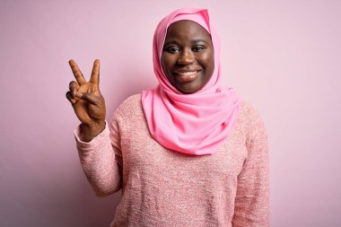 Genç Afro-Amerikan artı cüsseli kadın soyutlanmış pembe arka plan üzerine Müslüman başörtüsü takıyor ve iki numarayı işaret ederek kendinden emin ve mutlu bir şekilde gülümsüyor..