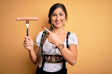 Geleneksel Ekim Festivali elbisesi giyen genç esmer kadın elinde sosisle elini ve parmağını göstererek mutlu mesut işaret ediyor.