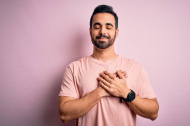 Sakallı, sıradan tişörtlü, pembe arka planda duran, göğsünde elleri kapalı, yüzünde minnet ifadesi olan genç yakışıklı bir adam. Sağlık kavramı.