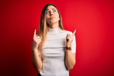 Mavi gözlü güzel sarışın kadın kırmızı arka planda günlük beyaz tişört giyiyor üzgün ve üzgün görünüyor, parmaklarıyla yön gösteriyor, mutsuz ve depresif..
