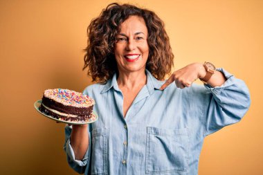 Orta yaşlı, güzel bir kadın, izole edilmiş sarı arka planda durup kendini işaret eden sürpriz yüzüyle pasta tutuyor.