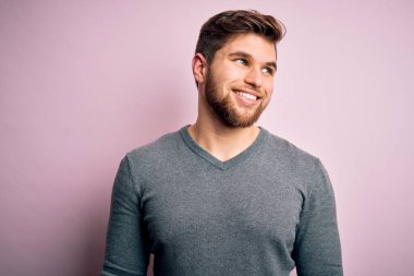 Sakallı, mavi gözlü, sıradan süveter giyen, yüzünde gülümseme, doğal bir ifade olan genç, yakışıklı sarışın bir adam. Kendinden emin gülen.