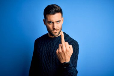 Young handsome man wearing casual sweater standing over isolated blue background Showing middle finger, impolite and rude fuck off expression