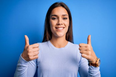 Genç, güzel esmer kadın mavi arka plan başarı tabelasının üzerinde dikilmiş gülümsüyor ve mutlu bir şekilde olumlu jest yapıyor. Neşeli ifade ve kazanan jest.