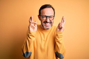 Orta yaşlı, beyaz bir adam, sarı arka planda gözlük ve süveter takıyor. Umut ve gözler kapalı bir şekilde gülümseyerek işaret ediyor. Şans ve batıl inanç..