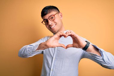 Sarı arka planda inek gözlüğü takan genç, yakışıklı İspanyol iş adamı. Sevgiyle gülümsüyor. Elleriyle kalp sembolü yapıyor. Romantik konsept.