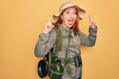 Sırt çantası ve sarı arka planda şapka takan genç kızıl saçlı kadın şaşırmış, şaşırmış ve parmaklarıyla işaret ediyor ve kollarını kaldırıyordu..