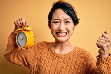 Young beautiful chinese woman holding vintage alarm clock over isolated yellow background annoyed and frustrated shouting with anger, crazy and yelling with raised hand, anger concept