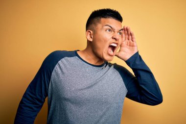 Sarı arka planda dikilmiş, elleri ağızda bağırarak bağıran gündelik tişört giyen genç, yakışıklı Latin adam. İletişim konsepti.