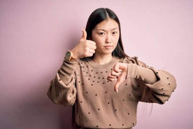 Genç, güzel Asyalı bir kadın pembe arka plan üzerinde şık bir süveter giyiyor. Baş parmak işareti yapıyor, anlaşmazlık ve uzlaşma ifadesi takınıyor. Çılgın çatışma