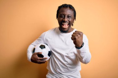 Genç Afrikalı Amerikalı oyuncu futbol topunu sarı arka planda tutarak oynuyor. Gururla bağırıyor, zaferi ve başarıyı kutluyor. Heyecanlı, coşkulu, coşkulu.