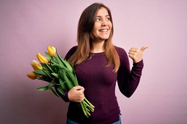Sarı lalelerden oluşan romantik buketi pembe arka planda tutan genç sarışın kadın gülümsüyor ve başparmağıyla yan tarafa bakıyor..