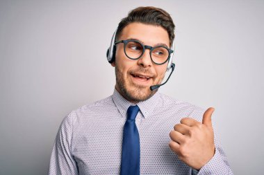 Genç çağrı merkezi operatörü mavi gözlü, gözlüklü, kulaklıklı, gülümseyen, gülümseyen ve baş parmağıyla yan tarafı gösteren iş adamı..