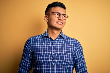 Sarı arka plan üzerine gündelik gömlek ve gözlük takan genç, yakışıklı Latin adam yüzünde gülümseme, doğal bir ifade ile yan yana bakıyor. Kendinden emin gülen.