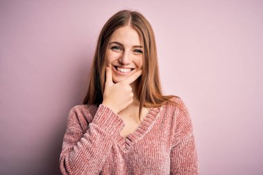 Genç, güzel, kızıl saçlı bir kadın pembe arka plan üzerine günlük süveter giyip kameraya bakarken kolları çapraz ve çenesi açık gülümsüyor. Olumlu düşünüyorum..