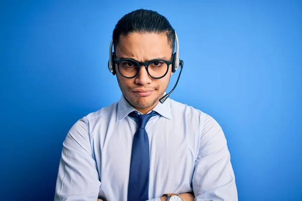 Young brazilian call center agent man wearing glasses and tie working using headset skeptic and nervous, disapproving expression on face with crossed arms. Negative person.