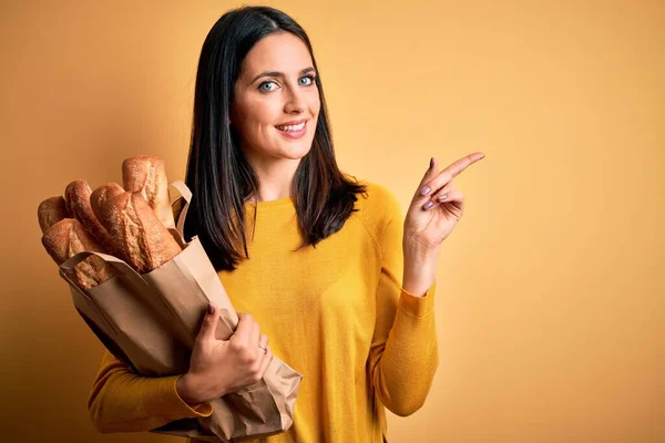 年轻的女人 蓝眼睛 手里拿着纸袋 上面夹着面包 背景是孤立的 黄色的 她非常高兴地把手指头指向旁边 — 图库照片