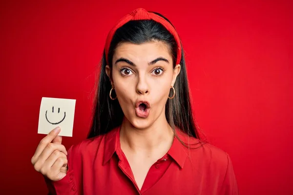 Young beautiful brunette woman holding reminder paper with smile emoji message scared in shock with a surprise face, afraid and excited with fear expression