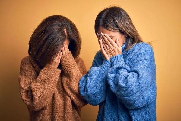 Middle age beautiful couple of sisters wearing casual sweater over isolated yellow background with sad expression covering face with hands while crying. Depression concept.