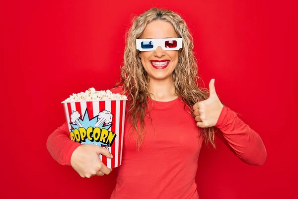 Young beautiful blonde woman watching movie using 3d glasses eating popcorn Smiling happy and positive, thumb up doing excellent and approval sign