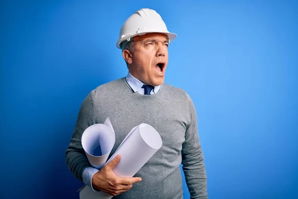 Middle age handsome grey-haired architect man wearing safety helmet holding blueprints angry and mad screaming frustrated and furious, shouting with anger. Rage and aggressive concept.