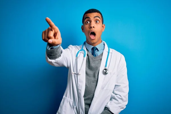 Handsome african american doctor man wearing coat and stethoscope over blue background Pointing with finger surprised ahead, open mouth amazed expression, something on the front