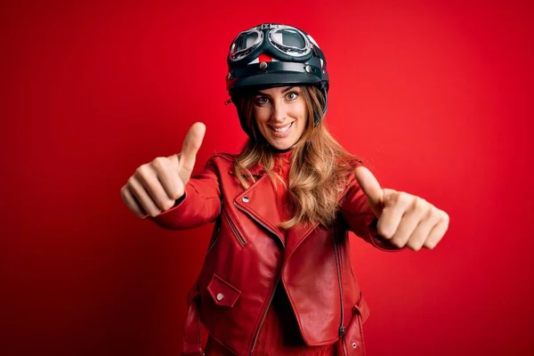 若い美しいブルネットのオートバイの女性は 手で肯定的なジェスチャーを行うことを承認赤い背景に元ヘルメットを身に着けています 親指アップ笑顔と成功のために幸せ 勝者のジェスチャー — ストック写真