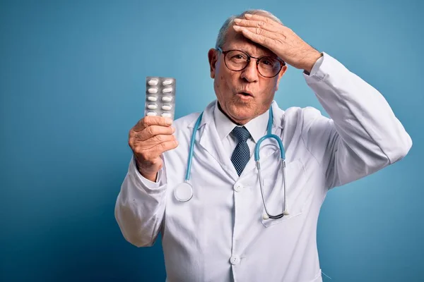 Senior grey haired doctor man holding pharmaceutical pills over blue background stressed with hand on head, shocked with shame and surprise face, angry and frustrated. Fear and upset for mistake.
