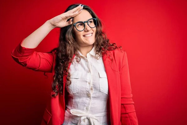 Young beautiful woman with curly hair wearing jacket and glasses over red background very happy and smiling looking far away with hand over head. Searching concept.