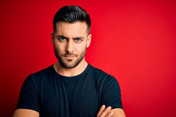 Young handsome man wearing casual black t-shirt standing over isolated red background skeptic and nervous, disapproving expression on face with crossed arms. Negative person.