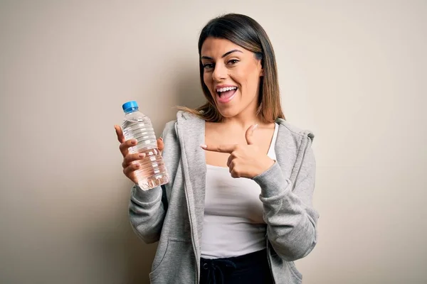 年轻美丽的黑发女运动员 手指手画脚地在与世隔绝的白色背景上喝着一瓶水 非常快乐 — 图库照片