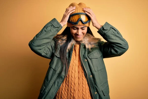 痛みや片頭痛のために絶望的な頭痛やストレスに苦しんで冬の天候のためのスキー眼鏡やコートを身に着けている若い美しいヒスパニック系の女性 手を頭に — ストック写真