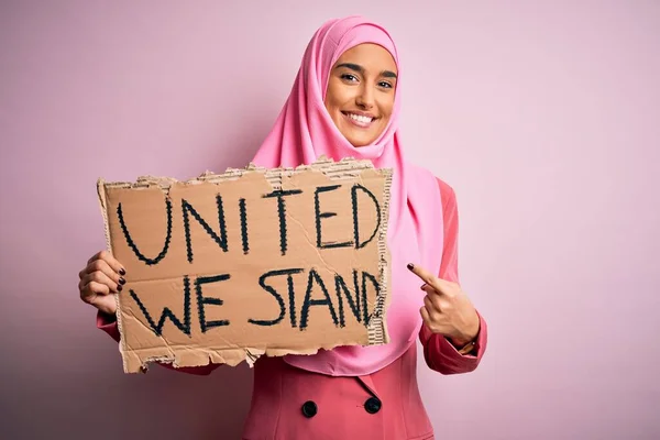 ピンクのイスラム教徒のヒジャーブを身に着けている若い活動家の女性が団結してバナーを保持我々は手と指で非常に幸せなポインティングメッセージを立つ — ストック写真