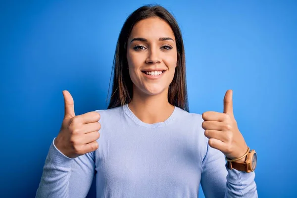 Young beautiful brunette woman wearing casual sweater standing over blue background success sign doing positive gesture with hand, thumbs up smiling and happy. Cheerful expression and winner gesture.