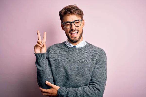 眼鏡をかけた髭を生やした若いハンサムな男とピンクの背景の上に立っているセーターは カメラで勝利のサインをして幸せそうな顔をして笑っています 第二番 — ストック写真