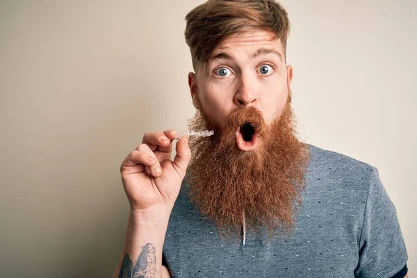 Irish redhead man with beard holding dental invisible aligner for tooth correction scared in shock with a surprise face, afraid and excited with fear expression