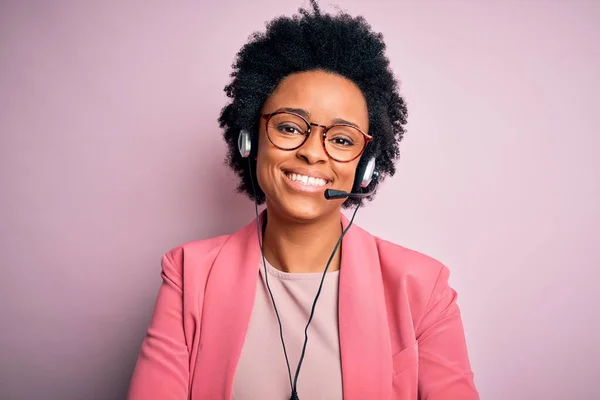 Young African American call center operator woman with curly hair using headset happy face smiling with crossed arms looking at the camera. Positive person.