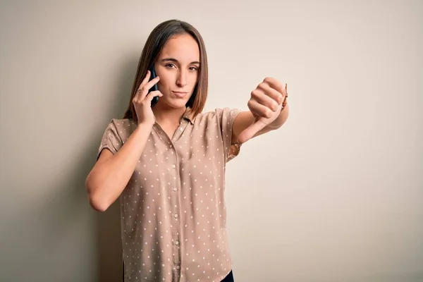 白い背景の上にスマートフォンで会話をしている若い美しい女性は 親指を下にジェスチャーで拒絶と否定を示す不満と怒りを見ています 悪い表現 — ストック写真