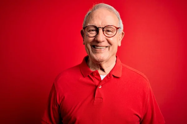 Grey haired senior man wearing glasses and casual t-shirt over red background with a happy and cool smile on face. Lucky person.
