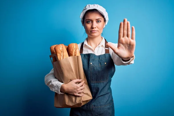 年轻美丽的面包师女人 蓝眼睛 手握纸袋 手拿着面包 手拿着面包 做手势 严肃而自信 做手势防守 — 图库照片