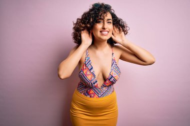 Genç, güzel bir Arap kadın, mayo ve pembe arka planda güneş gözlüğü takarak tatilde. İki elini de kulak hareketi yaparken duymaya çalışıyor. Dedikoduya meraklı. İşitme problemi, sağır