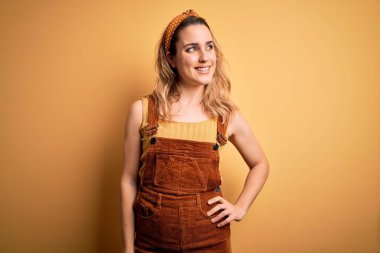 Tulum ve diadem giymiş genç, güzel sarışın kadın sarı arka planda dikilip yüzünde gülümseme, doğal bir ifade ile yan yana bakıyor. Kendinden emin gülen.