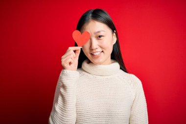 Genç Asyalı bir kadın kırmızı kalp kağıdını kırmızı izole edilmiş arka planda tutarken mutlu bir yüz, kendine güvenen bir gülümsemeyle dişlerini gösteriyor.