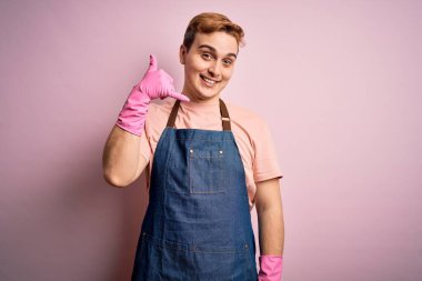 Genç, kızıl saçlı, ev işi yapan, önlüklü, eldivenli, gülümseyen, el ve parmaklarıyla telefonla konuşan genç bir temizlikçi. İletişim kavramları.