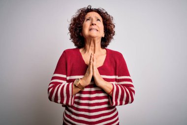 Orta yaşlı güzel kıvırcık saçlı kadın beyaz arka planda sıradan çizgili süveter giyiyor. Elleriyle yalvarıyor ve dua ediyor. Yüzünde umut ifadesi var. Çok duygusal ve endişeli. Yalvarıyorum..