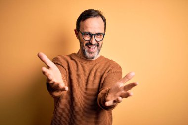 Orta yaşlı, beyaz, kahverengi süveter ve gözlük takan izole edilmiş sarı arka planda gülümseyen, yardım eli uzatan ve kabul gören bir adam..