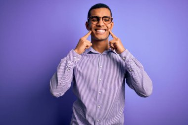 Yakışıklı Afro-Amerikan erkeği çizgili gömlek ve gözlüklerle mor arka planda gülümsüyor, parmakları açık ve neşeli gülümsemeye zorluyor.