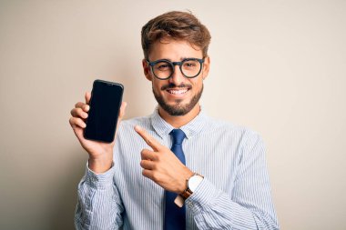 Akıllı telefonu elinde tutan genç bir işadamı izole edilmiş beyaz arka planda çok mutlu bir şekilde el ve parmağıyla işaret ediyor.