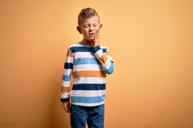 Küçük, beyaz, mavi gözlü, sarı arkaplan üzerine renkli çizgili gömlek giyen, diş ağrısı ya da diş ağrısı yüzünden ağıza acı dolu bir ifadeyle dokunan genç bir çocuk. Dişçi